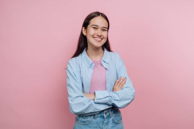 Genç bir kadın kollarını kavuşturup gülümseyerek kendine güvenerek ayakta duruyor. Pembe bir bluzun üzerine çizgili bir gömlek giyiyor ve parlak, pembe bir arka plana karşı poz veriyor, neşeli bir atmosfer sergiliyor..