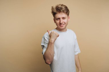 Sade beyaz tişörtlü genç bir adam bej bir arka planın önünde duruyor, gülümsüyor ve baş parmak işareti yapıyor. Neşeli ve arkadaş canlısı görünüyor. Olumlu bir hava yayıyor..