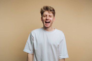 Genç bir adam mutluluğunu geniş bir gülümsemeyle ifade eder, gerçek bir neşe sergiler. Beyaz tişört giymiş, bej renginde yumuşak bir fonda duruyor..