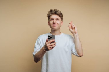 Genç bir adam sağ eliyle yukarıyı gösterip gülümserken sol elinde bir akıllı telefon tutuyor. Açık renkli bir odada gibi görünüyor, rahat ve pozitif bir atmosfer izlenimi veriyor..