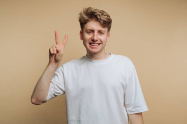 Saçı dağınık neşeli genç bir adam sağ eliyle barış işareti sergiliyor. Yumuşak, nötr bir arka planın önünde duruyor, pozitiflik ve sıcaklık yayıyor..