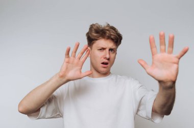 Genç bir adam düz bir arkaplan karşısında duruyor, elleri havada bir sürpriz ya da reddedilme hareketiyle. Yüzündeki ifade şoku ya da inanmazlığı yansıtıyor..