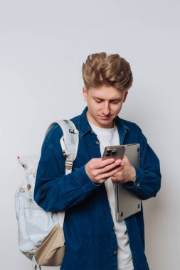 Genç bir adam içeride duruyor, diğer elinde bir tablet tutarken akıllı telefonuna odaklanmış. Mavi bir kot gömlek giyiyor ve sırtında bir sırt çantası asılı..