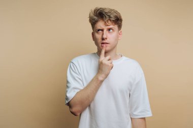 Genç bir adam düz bir bej duvarın karşısında duruyor. Yüzünde hafif bir sırıtma var ve çenesine dokunuyor, düşünceli bir tavır sergiliyor. Günlük kıyafet rahat bir stili yansıtıyor..