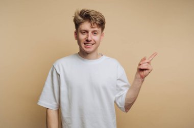 Basit beyaz tişört giyen genç bir adam açık bej bir arka fona sağ eliyle gülümsüyor ve işaret ediyor. Duruşu rahatlatıcı ve davetkar, pozitifliği gösteriyor..