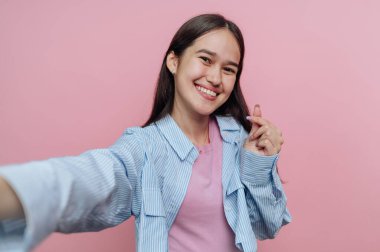 Neşeli genç bir kadın parlak pembe bir duvarın önünde duruyor, selfie çekerken geniş bir gülümseme sergiliyor. Çizgili bir gömlek ve pembe bir bluz giyiyor. Elleriyle oyuncu bir tabela sergiliyor..