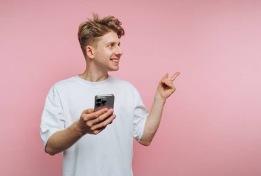 Saçları dağınık genç bir adam, canlı pembe bir arka plana karşı elinde bir akıllı telefon tutarak, coşkuyla gülümseyen ve işaret eden, günlük beyaz bir gömlekle kapalı bir yerde duruyor..