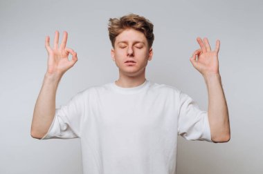 Gözleri kapalı bir genç adam, elleri meditasyon jesti ile rahat bir pozisyonda oturarak farkındalık yapar. İfadesi sükuneti yansıtıyor ve kapalı alanda minimalist bir alana odaklanıyor..