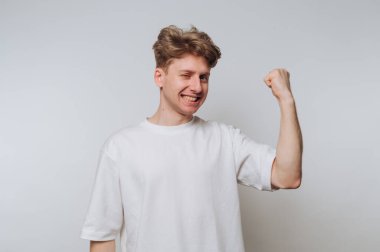 Açık kahverengi saçlı genç bir adam zaferle yumruğunu kaldırırken geniş bir şekilde gülümsüyor. Sıradan beyaz bir gömlek giyiyor ve düz bir zemine karşı duruyor..