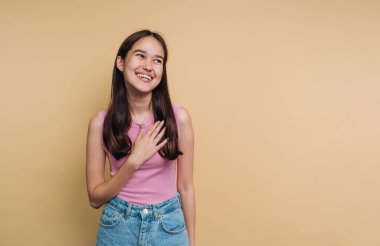 Neşeli genç bir kadın kendine güvenerek ayakta duruyor, gülümsüyor ve elini kalbine koyuyor, mutluluğu ve sıcaklığı iletiyor. Ayar basit bir sarı arkaplan içeriyor, neşeli ifadesini artırıyor..