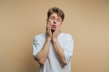 Genç bir adam, elleri yüzünde dinlenirken, gözle görülür şekilde stresli ya da hayal kırıklığına uğramış görünüyor. Renksiz bir arkaplanın önünde duruyor. Duygusal yorgunluk hissini iletiyor..