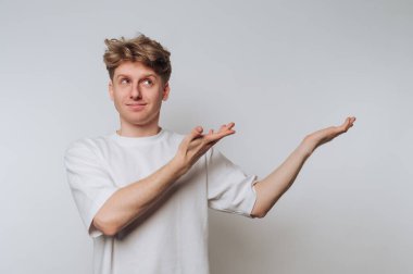 Sıradan beyaz tişörtlü genç bir adam düz bir iç mekanda duruyor. Kollarını dışa doğru uzatırken neşeli bir ifadesi var, sanki ilginç bir şey davet ediyor ya da sunuyor gibi..
