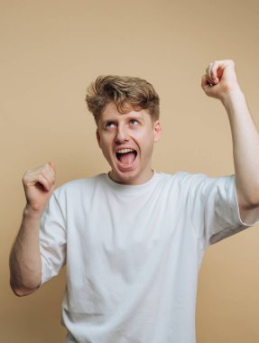 Açık kahverengi saçlı genç bir adam sevinçle kollarını kaldırıyor. Tarafsız bir arkaplanda coşkuyla gülümsüyor. Güçlü bir heyecan ve mutluluk hissi taşıyor..