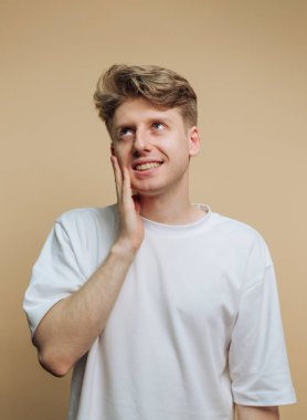 Açık renk saçlı ve sıradan beyaz gömlekli genç bir adam elini yüzünde dinlendirirken neşeyle gülümsüyor. Yumuşak bej arka plan sıcak bir atmosfer yaratır..