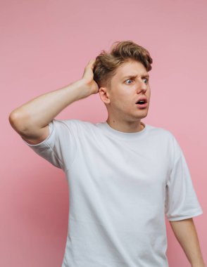Genç bir adam canlı pembe bir arka plana karşı duruyor, şaşkın bir bakış sergiliyor. Büyük gözlerle bakarken eli başının üzerinde duruyor, bir şok ya da şaşkınlık anı sergiliyor..