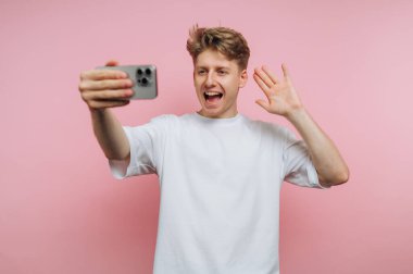 Açık kahverengi saçlı genç bir adam elini kaldırırken geniş bir gülümseme ile gülümsüyor. Önünde parlak pembe bir zemin üzerinde akıllı bir telefon tutuyor..