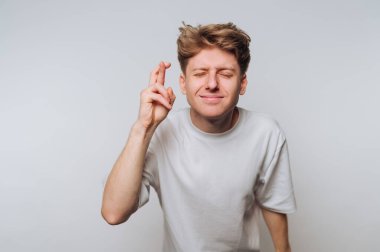 Genç bir adam, gözleri kapalı ve parmaklarını çapraz yaparak minimalist bir stüdyo ortamında umut veya iyi şans diliyor. Basit bir beyaz gömlek giyiyor..