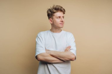 Kollarını kavuşturmuş, düşünceli bir şekilde yana bakan genç bir adam. O, basit beyaz bir tişört giydi. Tarafsız arka plan ifadesini vurguluyor..
