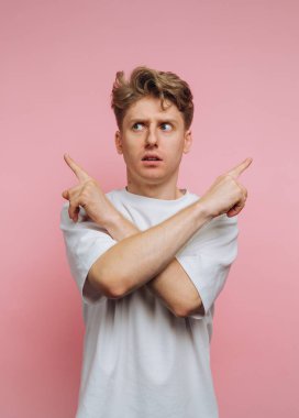Genç bir adam parlak pembe bir arka plana karşı duruyor, kollarını kavuşturup zıt yönleri işaret ederken şaşkın şaşkın bakıyor. Günlük kıyafetleri neşe katıyor..
