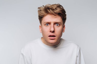 Genç bir adam şok olmuş bir bakış sergiliyor. Geniş gözleri ve hafif bir ağzı var. Tarafsız bir mekanda duruyor, basit bir arka planda, güçlü bir duygu aktarıyor..