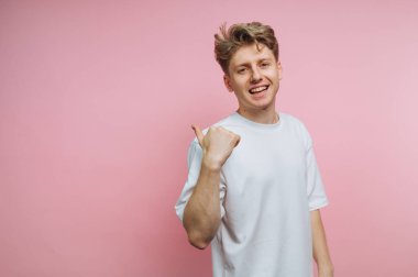 Genç bir adam başparmak işareti yaparken geniş bir gülümseme sergiliyor. Kısa, açık renkli saçları var ve sade beyaz bir tişört giyiyor. Arka plan yumuşak pembe bir renktir..