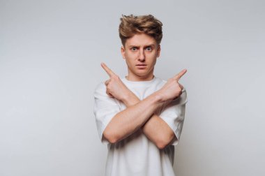 Saçları dağınık olan genç bir adam, parmaklarını çapraz kollarla iki farklı yönü işaret ederken şaşkın şaşkın bakıyor bir arka fonda duruyor. Onun ifadesi belirsizliği yansıtır..