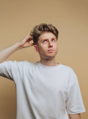 Düz beyaz tişört giyen genç bir adam derin düşüncelere dalmış görünüyor. Kafasını kaşıyarak yukarı bakıyor, düşüncelere dalmış gibi, yumuşak, nötr bir zemine doğru..