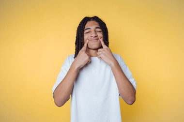 Saçları örgülü, gülümseyen ve sarı arka planda yanakları gösteren biri..