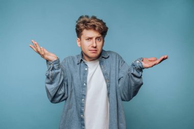 Kot gömlekli kafası karışmış genç bir adam mavi arka plana karşı omuz silkiyor..