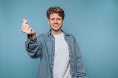 Sıradan kot gömlekli genç adam mavi arka planda parmaklarıyla kalp hareketi yapıyor..