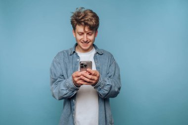 Mavi arka planda akıllı telefon kullanan genç adam gülümsüyor..