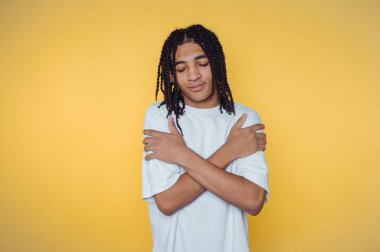 Saçları örgülü genç bir adam sarı arka planda kendine sarılıyor..