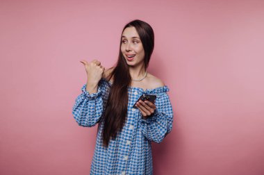 Mavi kareli elbiseli genç kadın akıllı telefon tutuyor, başparmağıyla omzunun üstünde işaret ediyor, gülümsüyor, pembe arka planda..