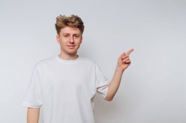 Beyaz tişörtlü genç adam düz bir arkaplanı işaret ediyor..
