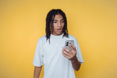 Saçları örgülü genç adam sarı arka planda akıllı telefona şaşırmış görünüyor..