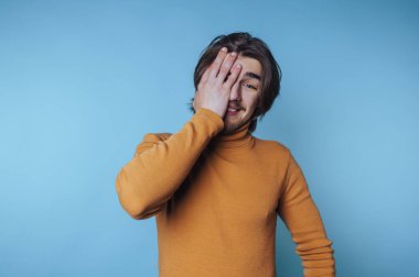 Turuncu kazaklı genç bir adam bir gözünü mavi arka planda tutuyor..