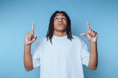 Saçları örgülü, beyaz tişörtlü genç bir adam mavi bir arka planı işaret ediyor..