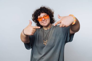 Kıvırcık saçlı, turuncu güneş gözlüğü takan ve günlük giysiler giyen, iki elini de gösteren, arka planda gülümseyen biri..