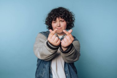 Kıvırcık saçlı biri mavi arka planda parmak kalp hareketi yapıyor..