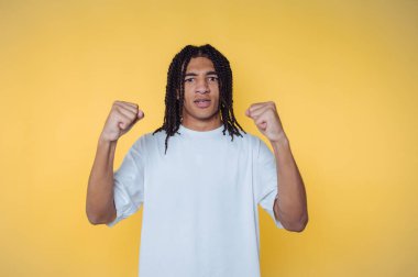 Saçlarını örmüş genç bir adam sarı arka planda kendine güvenen bir duruşta yumruklarını kaldırıyor..