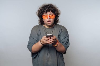 Kıvırcık saçlı ve elinde akıllı telefon tutan turuncu güneş gözlüklü şaşırmış bir insan..