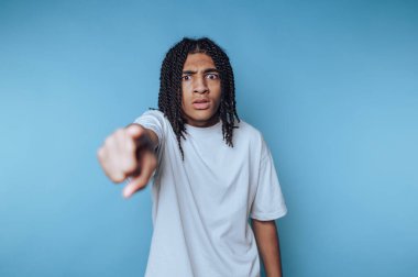 Örgülü saçlı, beyaz tişörtlü genç bir adam, mavi arka planda duruyor. Şaşırmış bir ifadeye sahipler ve ileriye bakıyorlar..