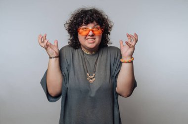 Kıvırcık saçlı, turuncu güneş gözlüklü ve gri gömlekli, elleri havada, düz bir arkaplan üzerinde..