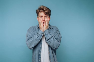 Mavi arka planda şok olmuş kot gömlekli genç adam..