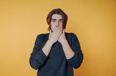 Uzun saçlı genç adam sarı arka planda ağzını kapatıyor..