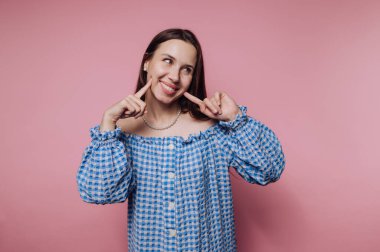 Mavi kareli elbiseli genç kadın pembe arka planda şakacı poz veriyor..