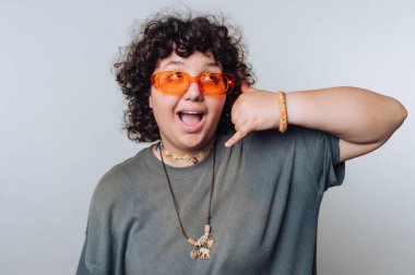 Kıvırcık saçlı, turuncu güneş gözlüğü ve mücevher takan, telefonla el hareketi yapan biri..