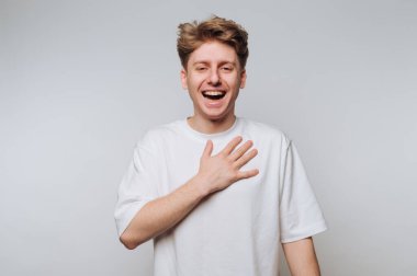 Genç adam elini göğsüne koyup gülüyor, beyaz tişört giyiyor.