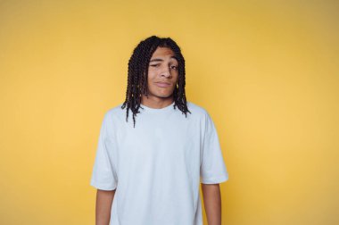 Saçları örgülü genç bir adam sarı bir arka plana göz kırpıyor..