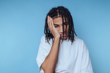 Saçları örgülü genç adam mavi arka planda yorgun görünüyor..
